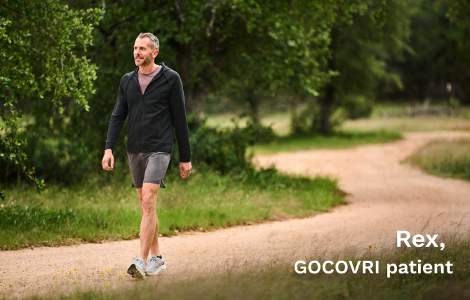Rex, a GOCOVRI patient, is taking a steady walk on a winding dirt path, surrounded by tall green trees. 